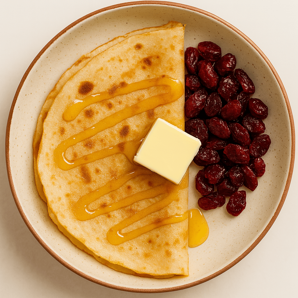 Breakfast plate with a folded crêpe topped with butter and honey, served with dried cranberries in a ceramic bowl.